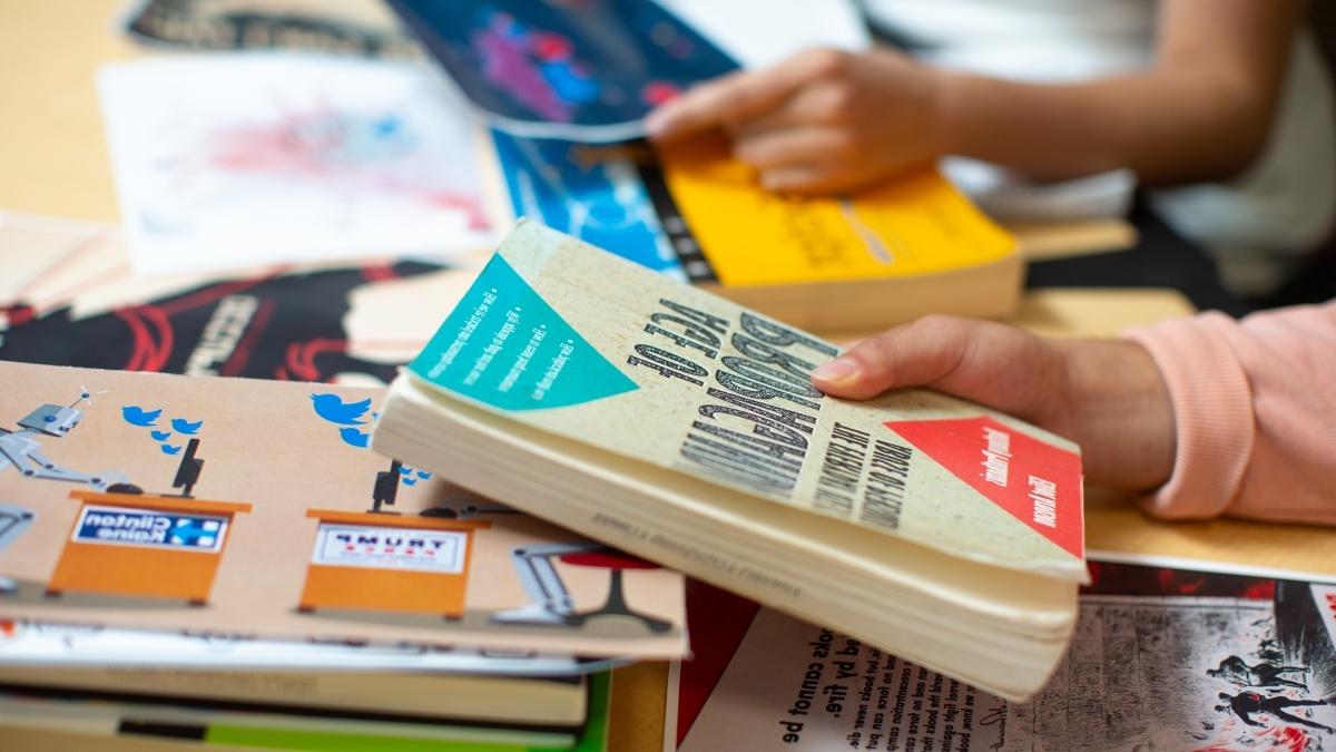 Student hand holding a textbook with other books piled underneath